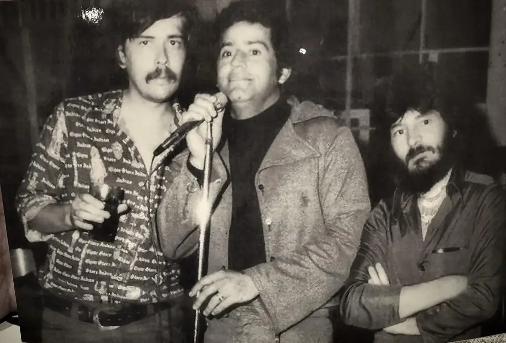 Fotografía en blanco y negro de tres músicos costarricenses en los años 80, posando con micrófono y copa en mano.