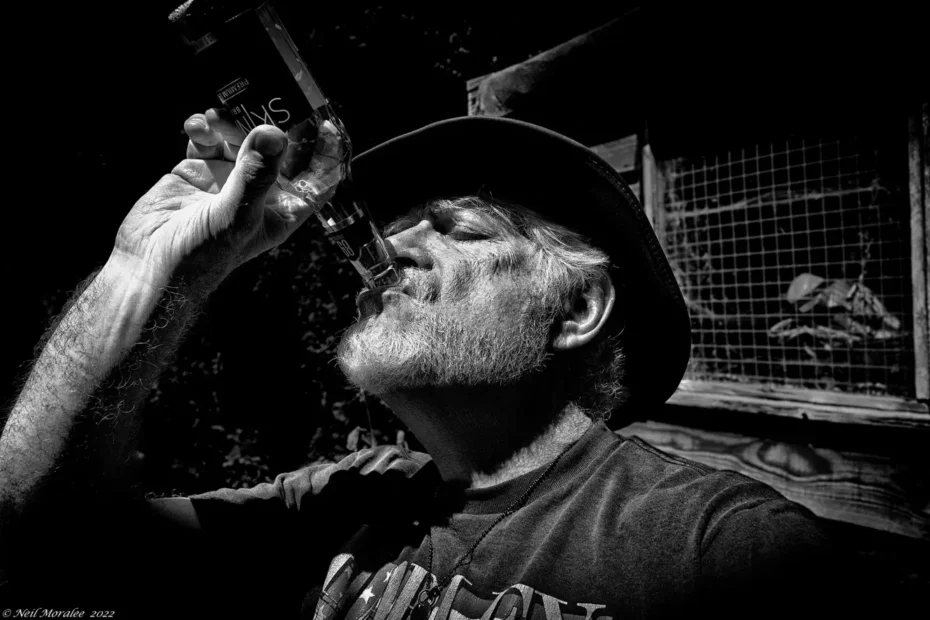 Hombre mayor con sombrero bebiendo de una botella de licor, retrato en blanco y negro.