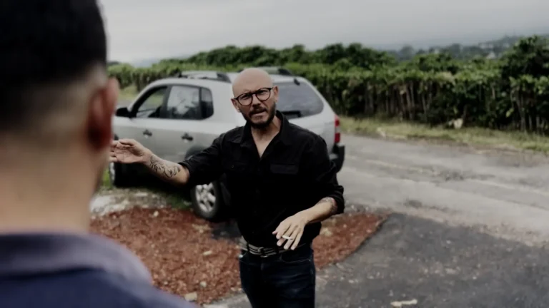 Hombre calvo con gafas y tatuajes dando indicaciones frente a un carro.