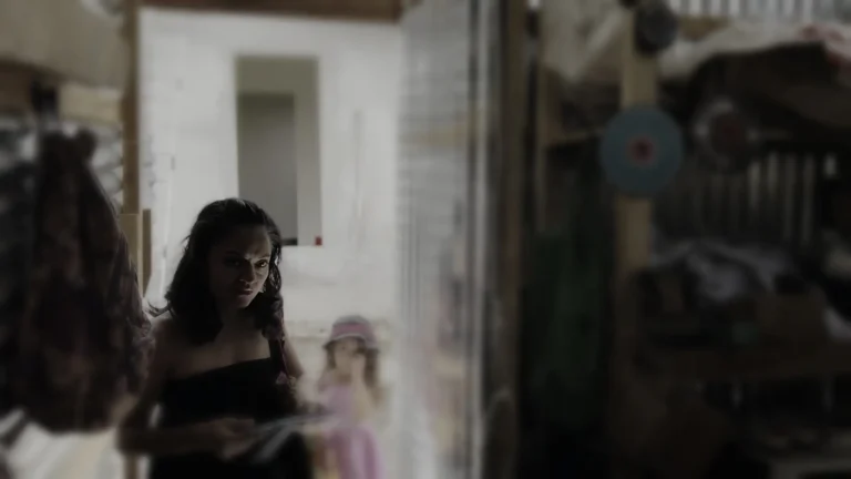 Mujer de cabello oscuro en primer plano, mirada seria, con una niña al fondo.