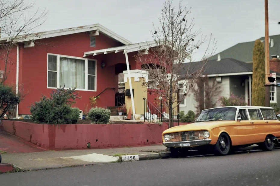 Casa roja de fachada sencilla con un carro clásico amarillo estacionado al frente, en una calle tranquila.