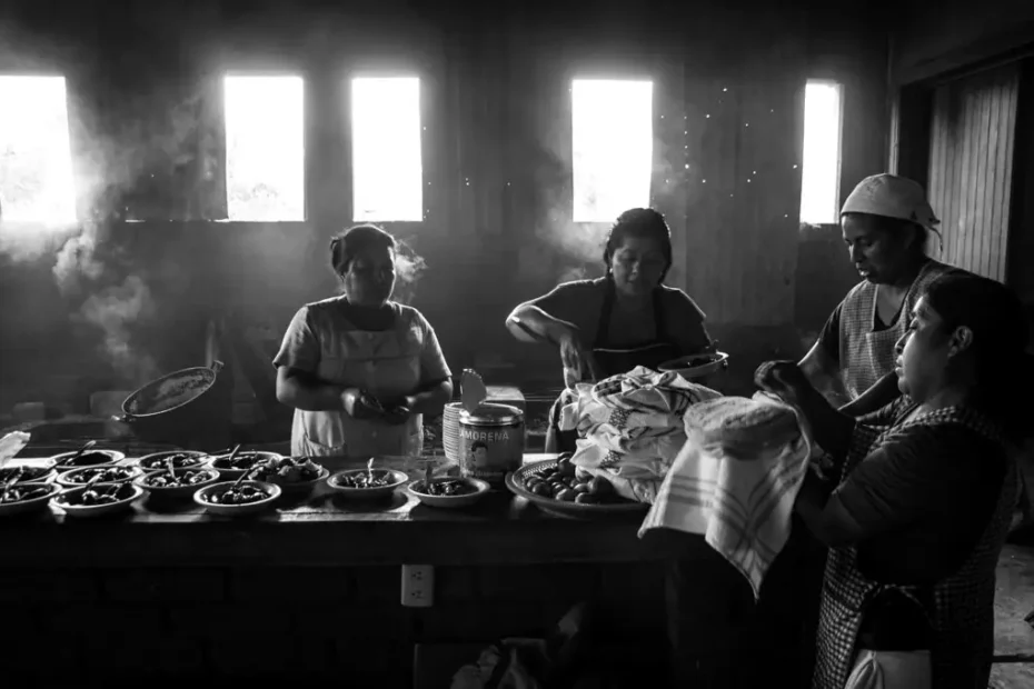 Grupo de mujeres cocinando en una cocina tradicional mexicana con humo y luz natural entrando por las ventanas.