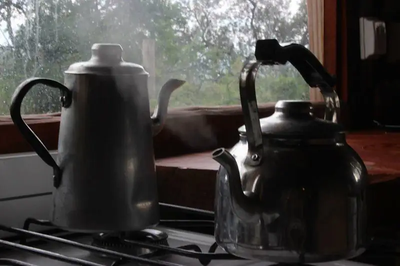 Dos pavas metálicas soltando vapor sobre una cocina encendida, con una ventana al fondo.