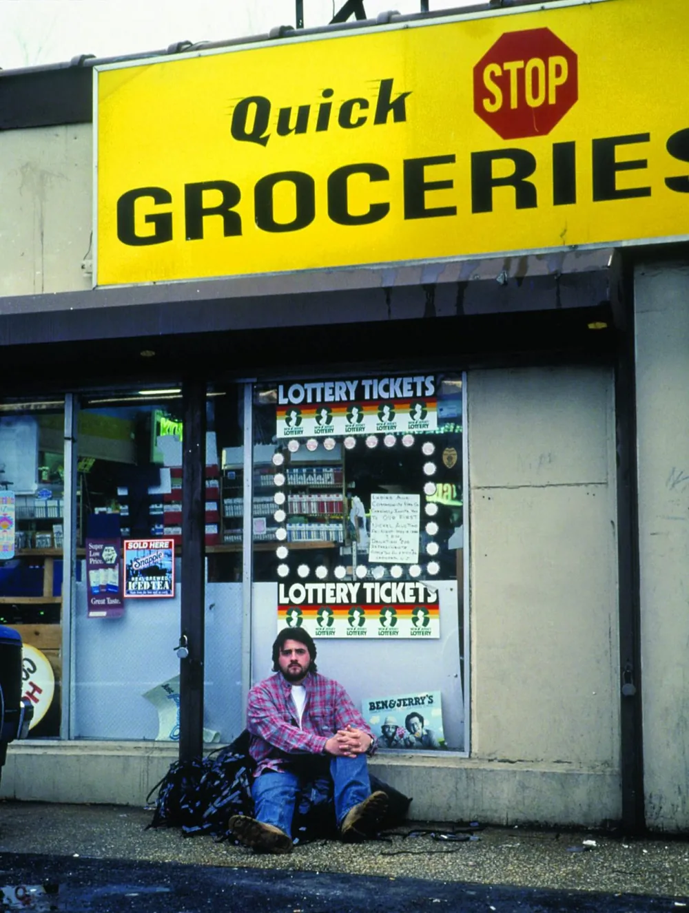 Kevin Smith sentado en la acera frente al Quick Stop Groceries, el mini súper donde se filmó Clerks.