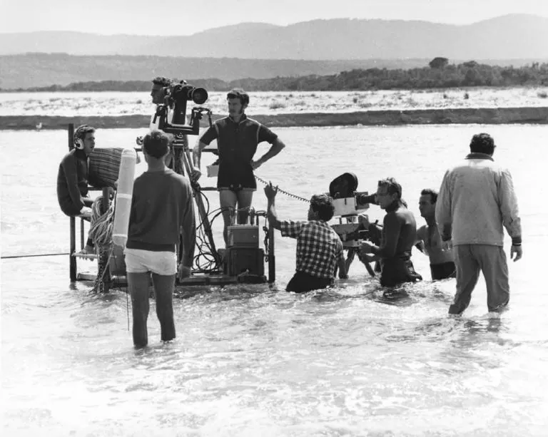 Equipo de rodaje con el agua a las rodillas, empujando una plataforma con la cámara.